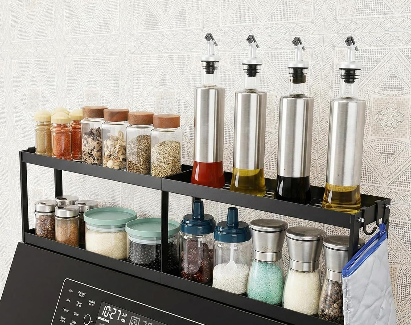 A black metal spice rack organizer mounted on a kitchen appliance, holding multiple glass jars with lids and four stainless steel oil bottles with pour spouts, against a light patterned wall.