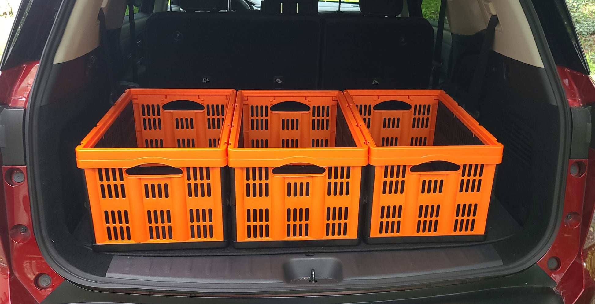 Four bright orange plastic utility cargo crates arranged side-by-side inside the open trunk of a red vehicle, with an outdoor grassy background visible through the rear hatch.