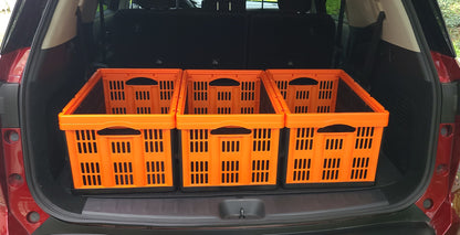 Four bright orange plastic utility cargo crates arranged side-by-side inside the open trunk of a red vehicle, with an outdoor grassy background visible through the rear hatch.