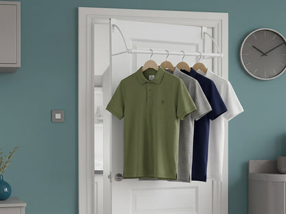 A green short-sleeve polo shirt hanging on a white over-the-door hanger in a doorway, with gray, navy, and white shirts behind it. The room has a blue wall, a round clock, and a light switch.