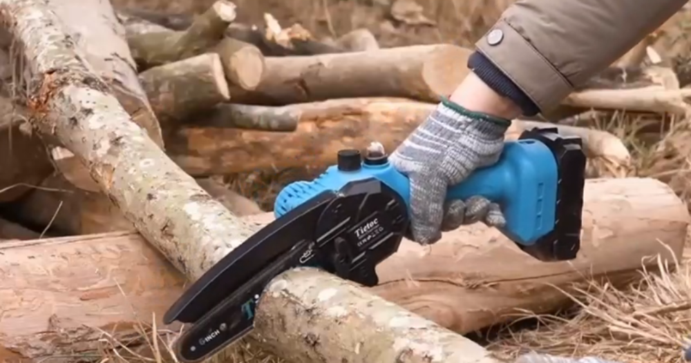 A person wearing a gray knit work glove holds a blue and black cordless mini chainsaw against a log outdoors.
